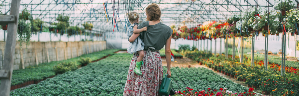 woman in skirt holding child on her left hip, standing in large community garden holding a watering can