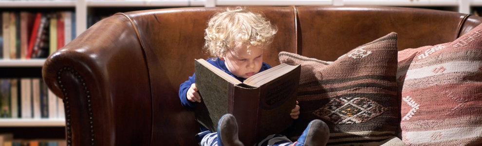 toddler reading large book on brown leather couch