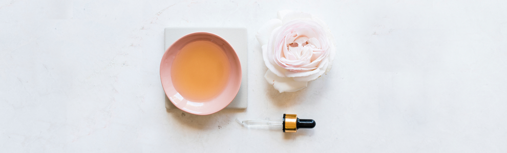 rose water in a pink cup, a light pink rose and a Pasteur pipette laid out on white surface